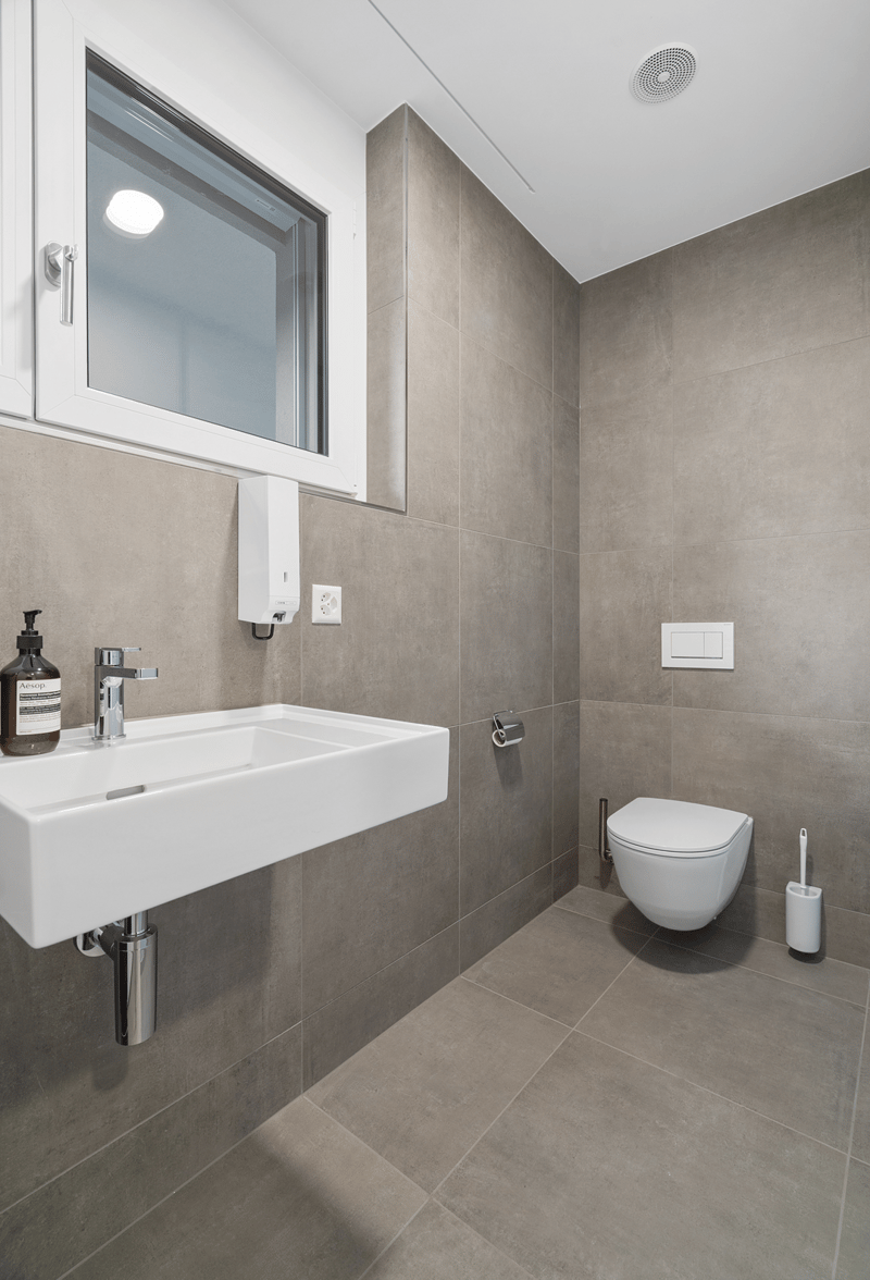 Small bathroom with white tiles, sink, toilet, toilet paper holder, soap dispenser, and vent.