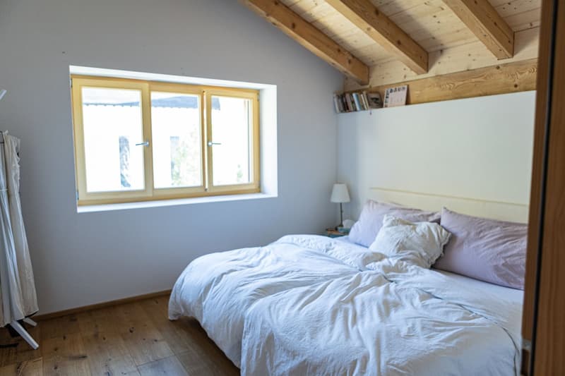 bedroom with wooden floor, white walls, bed with white sheets and purple pillows, wooden exposed beams, window with white frame