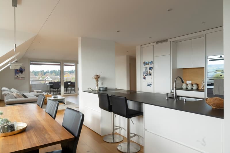 Contemporary kitchen with dining area, white cabinets, black countertops, stainless steel faucet, two barstools, large windows, living room with couch and coffee table