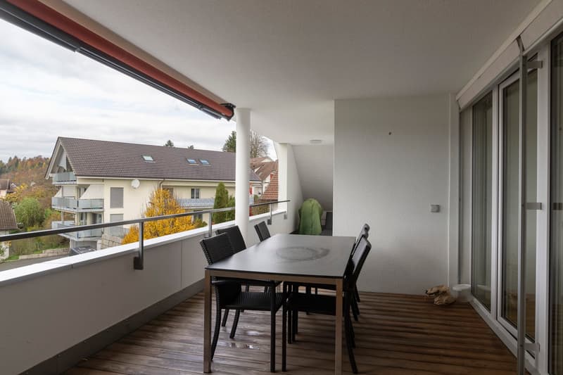 An outdoor wooden floor terrace with a round table and chairs, fenced with a metal railing. Behind the terrace there is a building with a brown roof and a view of the neighborhood.