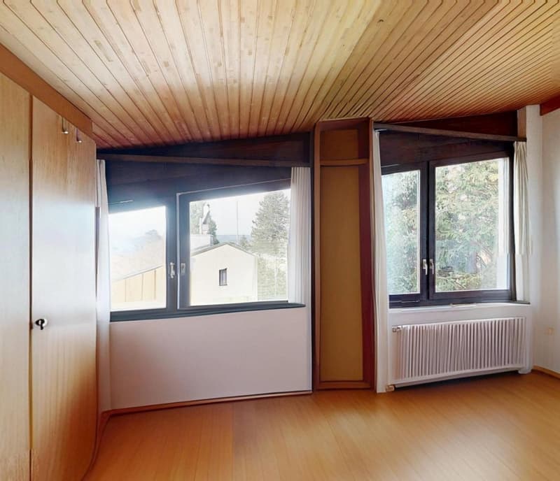 Wooden floor, wooden ceiling, two large windows, white radiator, open door, bright view of trees