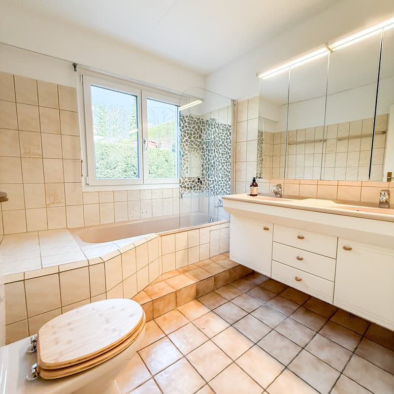 Bathroom: bathtub, double sink, toilet, tiled walls, tiled floor, large mirror, lighting, window
