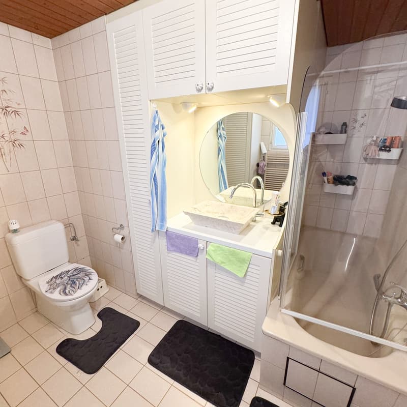 bathroom with toilet, sink, mirror, bathtub with glass door, tiled floor, towel hanging on a bar, and storage shelves