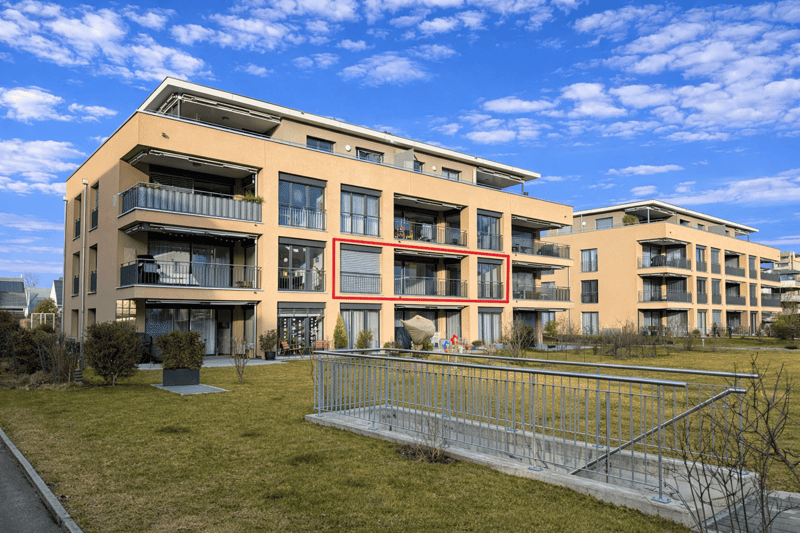 Modern apartment building with multiple balconies, beige exterior, large windows, and a landscaped front yard.