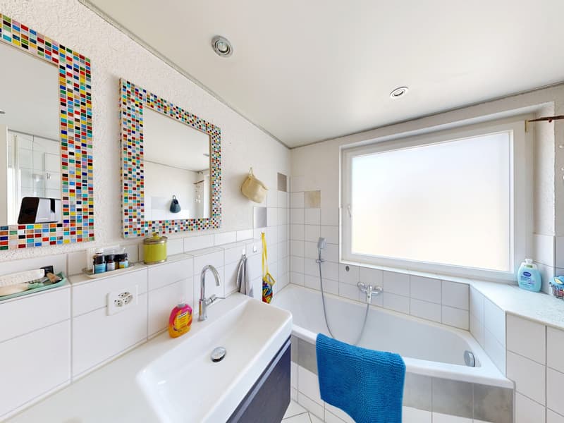 white tiles, bathtub, sink, two mirrors, window, blue towel, toiletries