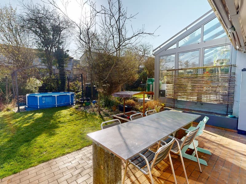 Outdoor dining table with chairs, brick patio, swimming pool, glass conservatory, trees, plants, and a fence