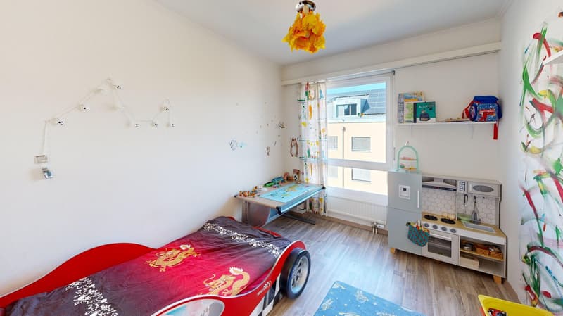 A child's bedroom with a red car bed, a desk, a toy kitchen, and a window with curtains.