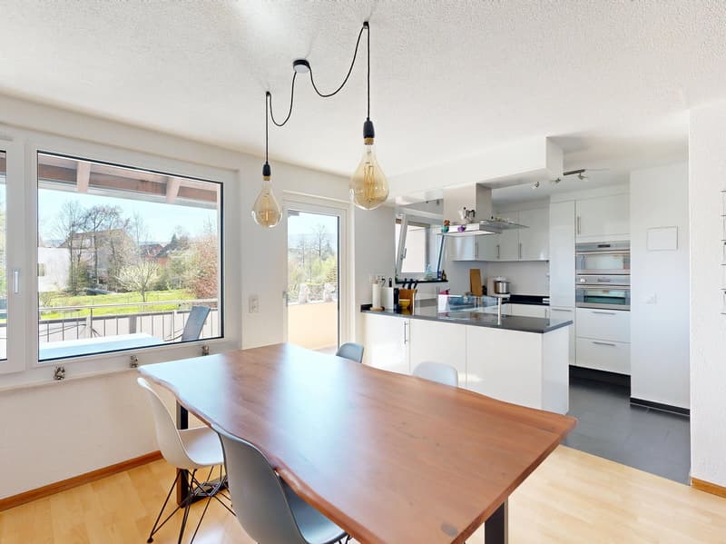 Dining room with wooden table, white chairs, large windows, balcony, modern kitchen with white cabinets, black countertops, and stainless steel appliances