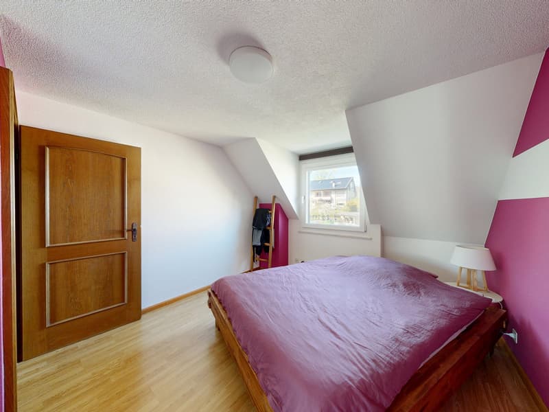 Bedroom with wooden floor, white walls, wooden door, bed with purple sheets, small table with lamp, window with view outside, clothes rack