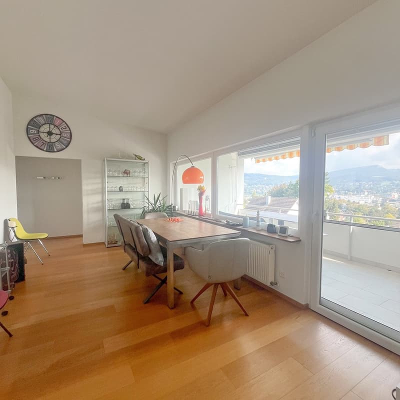 Dining room with wooden floor, large windows, wooden table, chairs, lamp, and decorative objects, balcony with railing, and distant view of trees and houses.