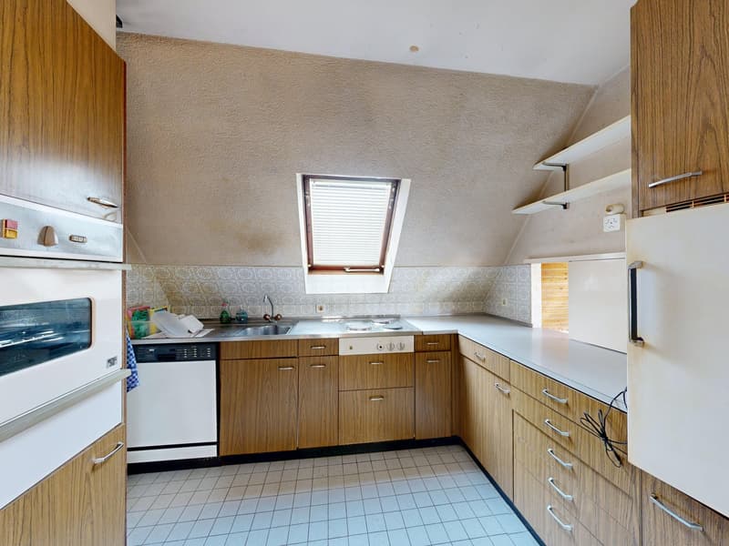 A kitchen with wooden cabinets, white appliances, and a white tiled floor. The kitchen includes a white oven, sink, faucet, dishwasher, gas stove, and refrigerator. There is a window with blinds and a roll of paper towels on the counter. There are also bo