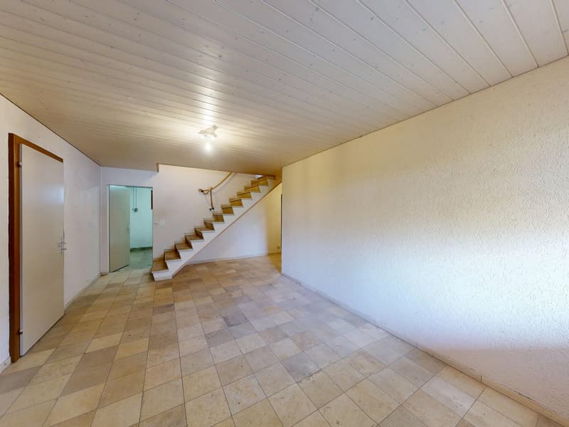 Empty room with tiled floor, white walls, and a wooden door. A staircase is visible in the background.