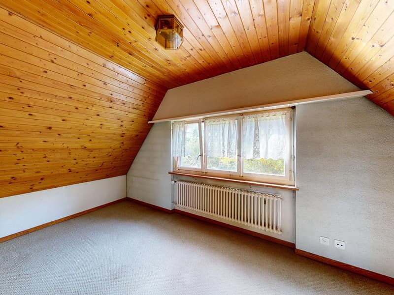 An empty attic room with wooden walls and ceiling, a white radiator, and a window with white curtains.