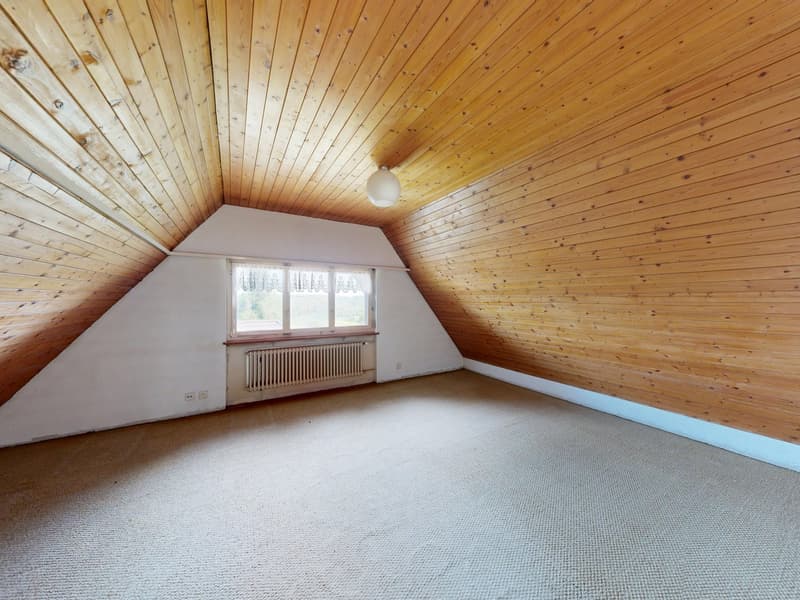 An empty attic room with wooden walls and ceiling, a window with white curtains, a radiator, and a ceiling lamp.