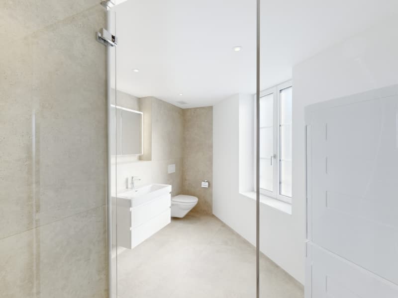 Spacious bathroom with white walls, large mirror, vanity sink, and toilet. The room has a clean, modern design with plenty of natural light from the window.
