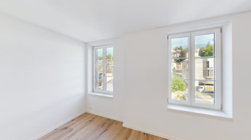 Spacious room with large windows, hardwood floors, and white walls. The room appears to have a balcony or terrace visible through the windows.