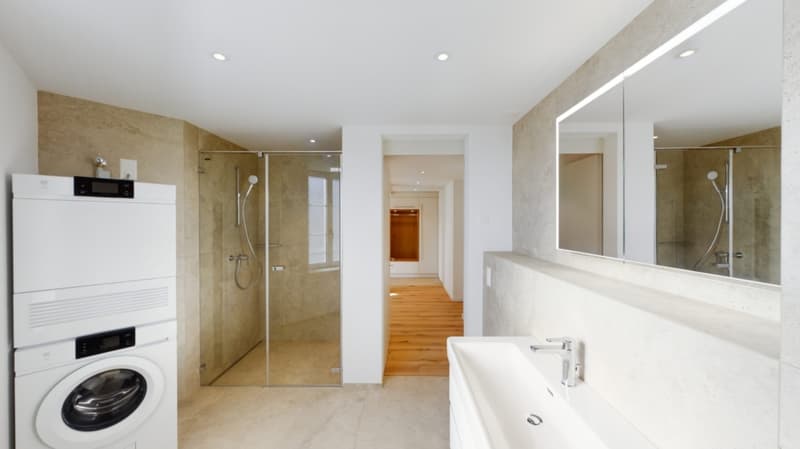 Large bathroom with beige tile walls, glass shower enclosure, vanity with sink, and a washing machine and dishwasher visible.