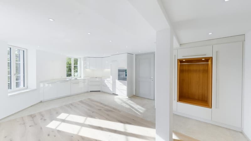 Spacious open-concept kitchen with white cabinets, stainless steel appliances, and hardwood flooring. Large windows provide ample natural light. Dishwasher is visible as one of the kitchen appliances.