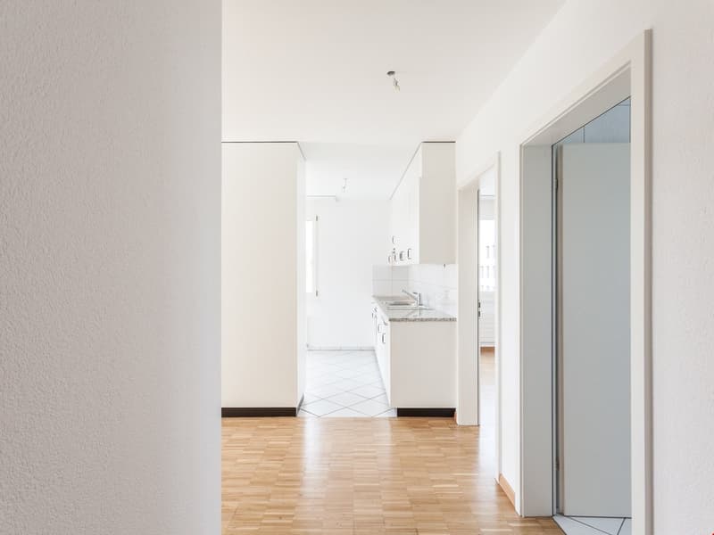 Empty room with wooden flooring, white walls, kitchen in the background, modern design, door on the right side