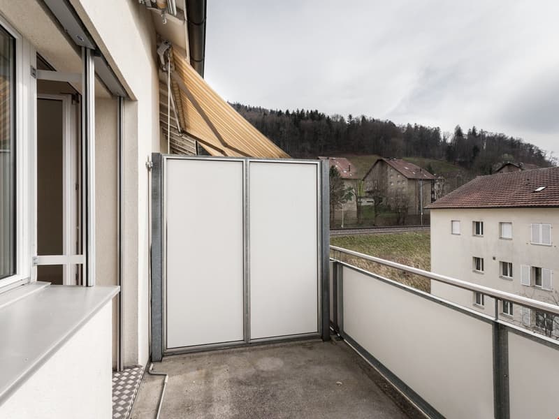 Closed balcony with white sliding door, metal railings, white wall with a large window, grey tiles on floor, fenced from outside.