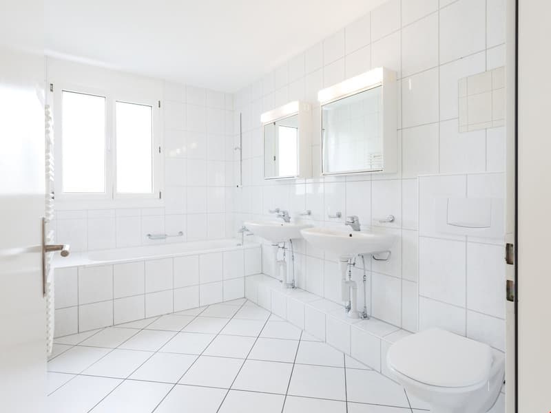 White tiled walls and floor, double sink, mirror, toilet, bathtub, natural light from windows