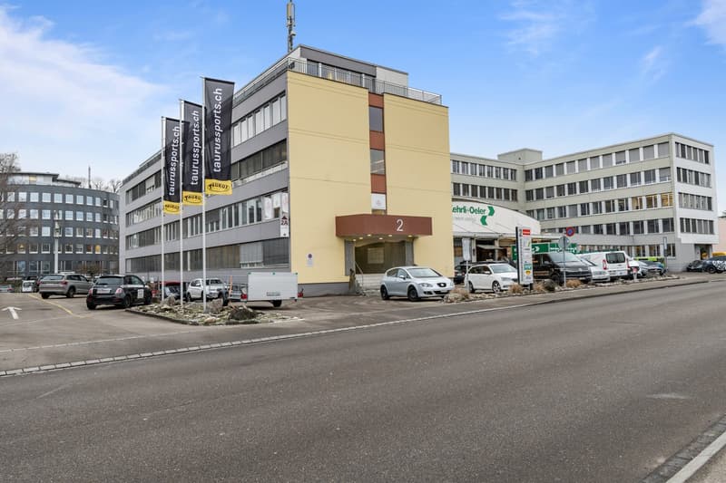 4-story building with yellow wall, glass windows, glass door, multiple flags outside, cars parked in front, and trees in the distance