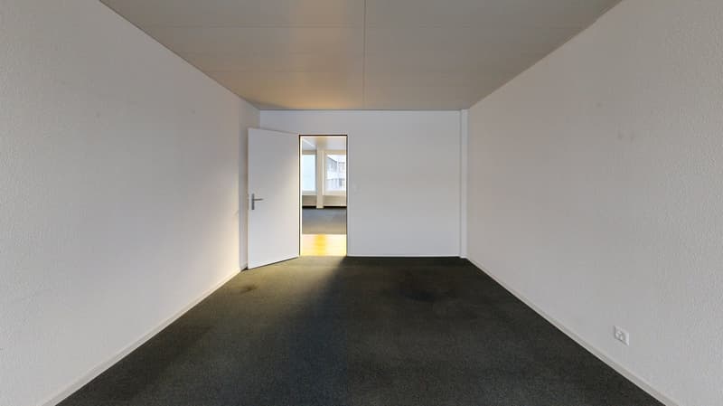 an empty room with white walls, a gray carpet, an open door, and a power outlet on the wall