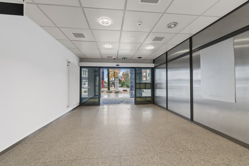 The image shows a commercial office space with a tiled floor, suspended ceiling with recessed lighting, and large glass windows that provide a view of the outdoor area. The space appears to be empty and ready for use.