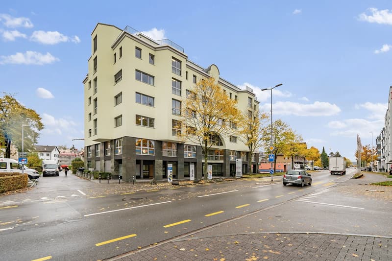 5-story apartment building with a modern, angular design, yellow exterior, and large windows. The building has balconies on the upper floors and is surrounded by trees with autumn foliage. There are cars parked on the street in front of the building and p