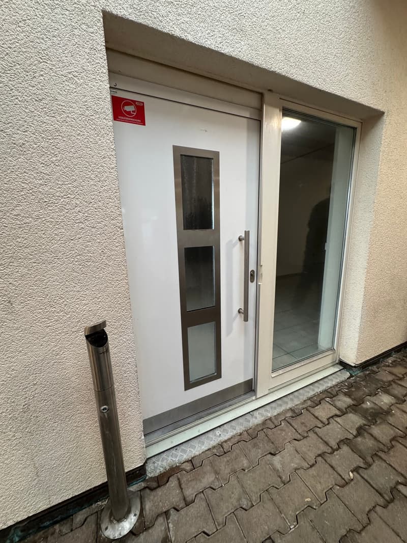 White entrance door with glass panels, metal handle, and a surveillance camera sign above it. The door is open, revealing a tiled floor inside.