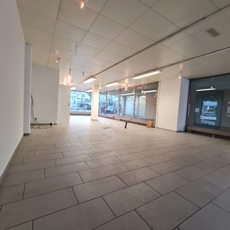 Empty commercial space with tile flooring, white walls, and fluorescent ceiling lights. The room also has windows and a broom leaning against the wall.