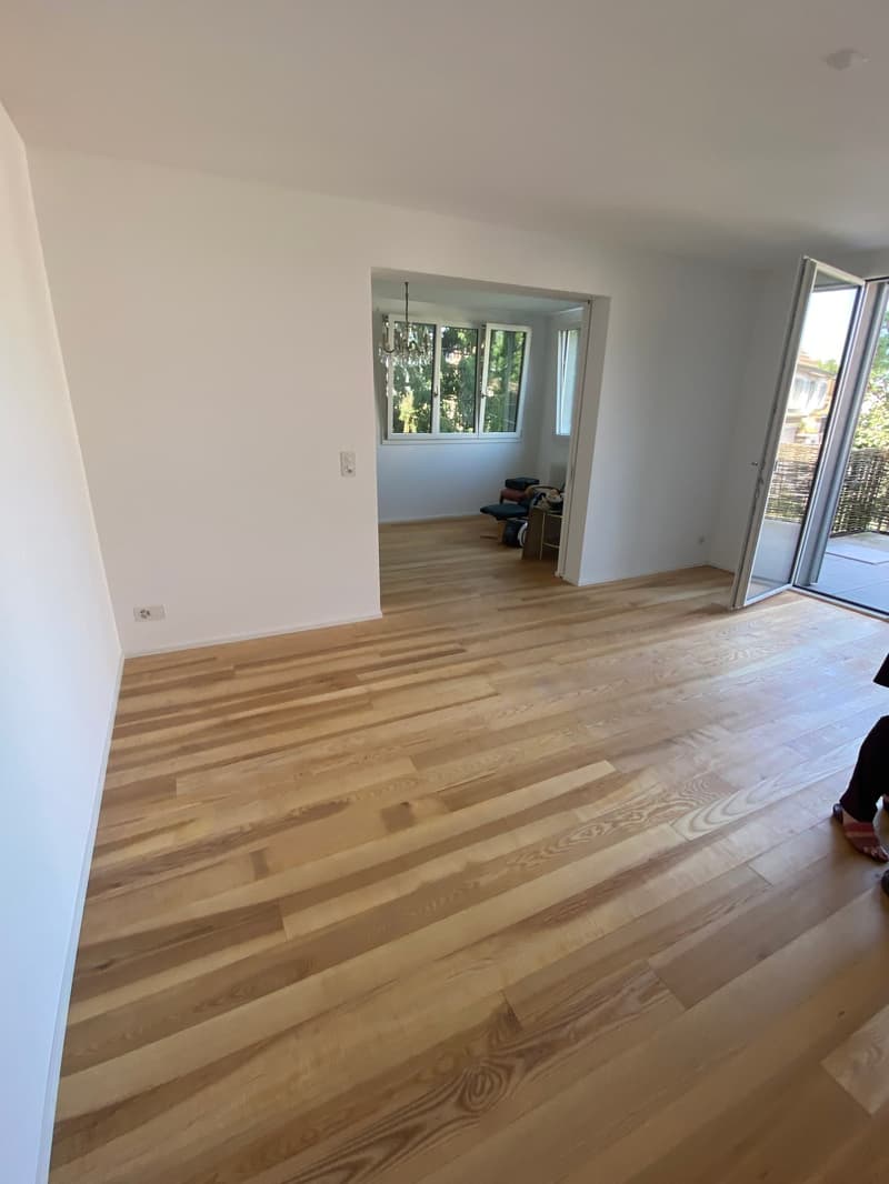 Spacious living room with hardwood flooring, white walls, and large windows providing natural light. The room has an open layout with a view of an adjacent room through the doorway.