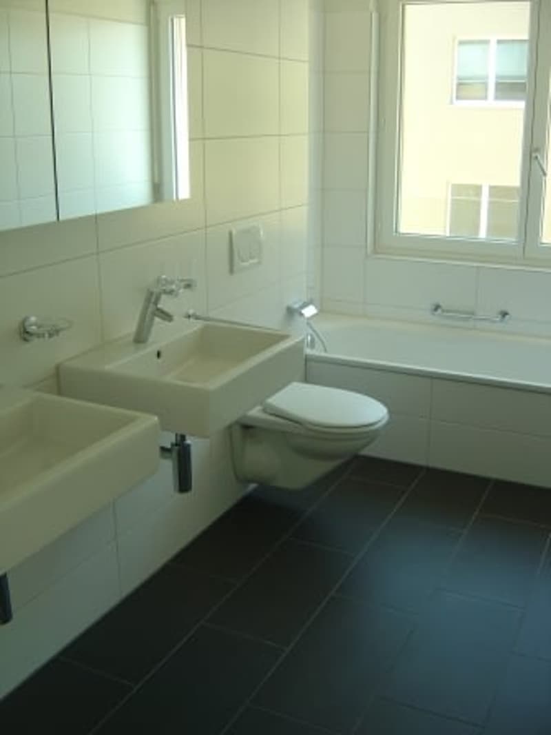 A contemporary bathroom with white ceramic tiles, a white bathtub, and double sinks with built-in faucets. A mirror, wall-mounted toilet, and natural light from the window.