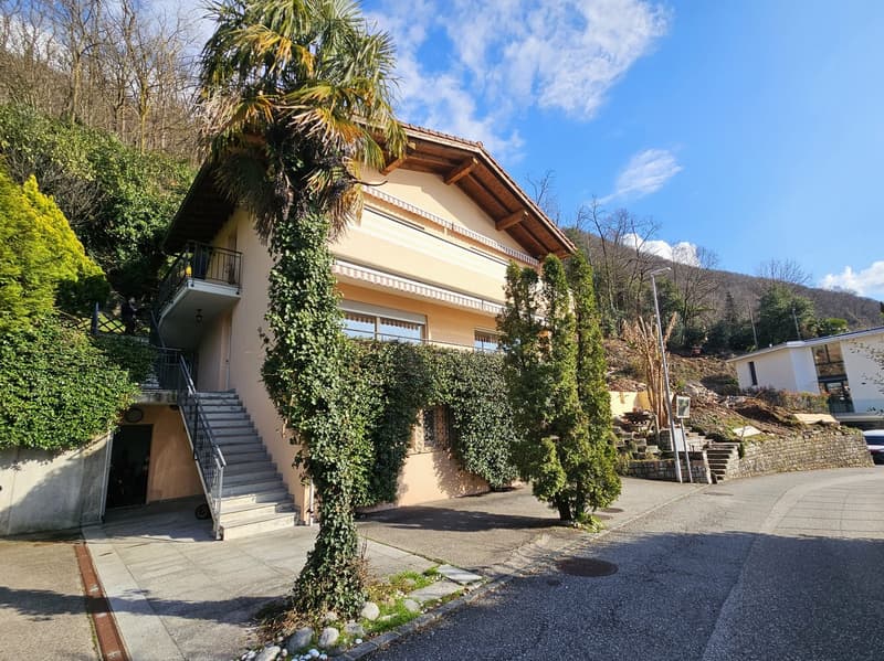 2-story house with a Mediterranean-style architecture, covered in ivy, surrounded by palm trees and greenery, with a balcony on the upper level and stairs leading up to the entrance.