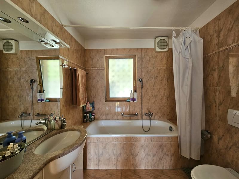 Large bathroom with marble-like tile walls, double vanity sink, bathtub, and shower curtain. There are also air conditioning units and storage shelves visible.