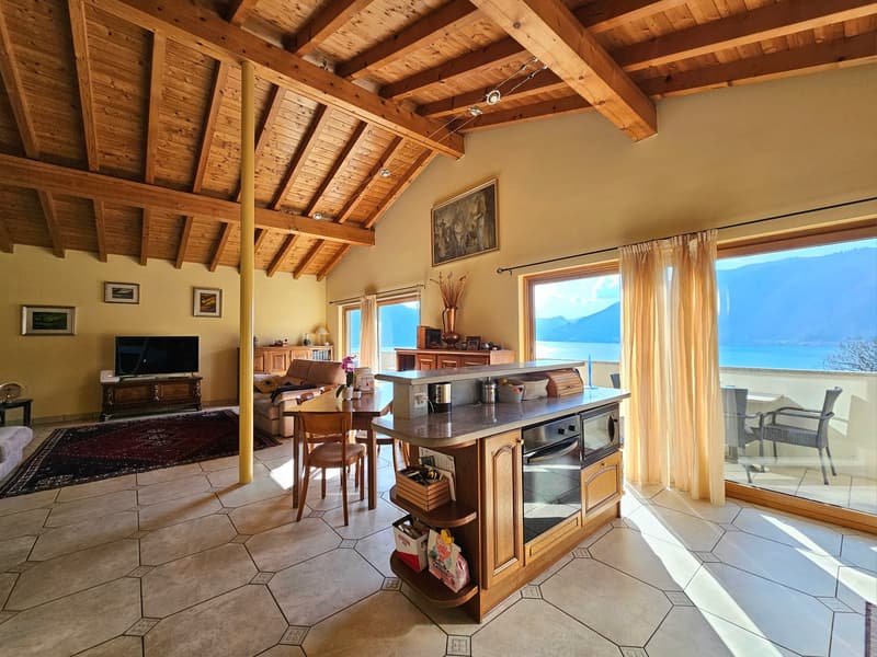 This image shows an open-concept living room and kitchen area with a vaulted wooden ceiling. The room has large windows that provide a scenic view of the surrounding landscape. The kitchen features a central island with storage and appliances, and the liv