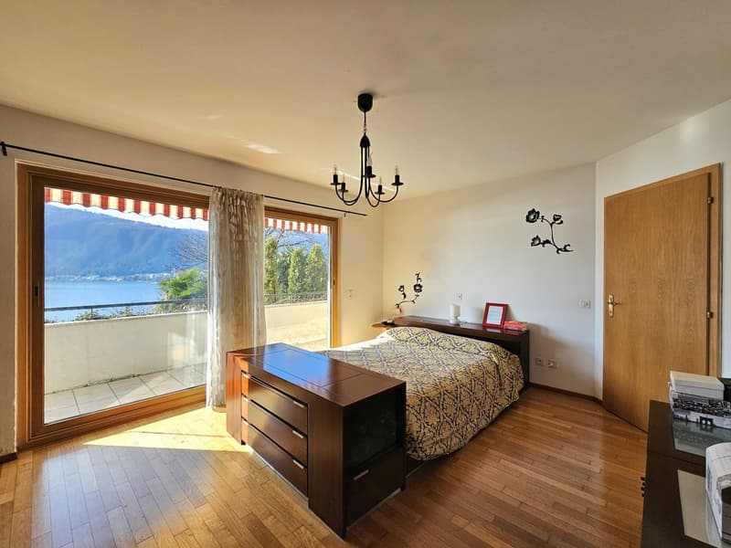 Spacious bedroom with hardwood floors, large windows overlooking a lake or body of water, a wooden dresser, and a chandelier lighting fixture. The room has a balcony or terrace with a view of the scenic outdoor landscape.
