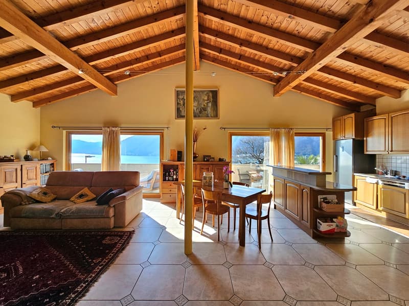 This image shows an open-concept living room and kitchen area with a vaulted wooden ceiling. The room has large windows that provide a scenic view of the mountains and lake outside. The living room features a comfortable brown sofa, while the kitchen area