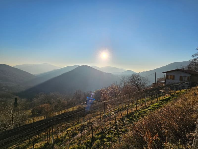 The image shows a scenic mountainous landscape with rolling hills, forests, and a small house or building visible on the hillside. The sun is shining through the hazy atmosphere, creating a warm, golden glow over the scene. The landscape appears to be in