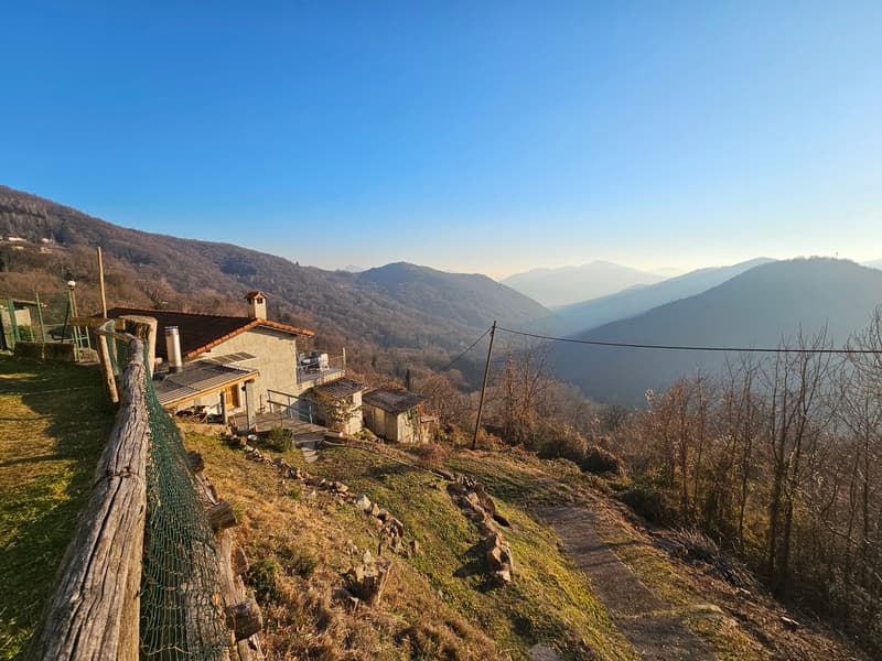 A rustic house situated on a hillside with a stunning view of the surrounding mountains and valleys. The house has a stone exterior and a chimney, surrounded by a grassy area and a wooden fence. The landscape features rolling hills, forests, and a clear b