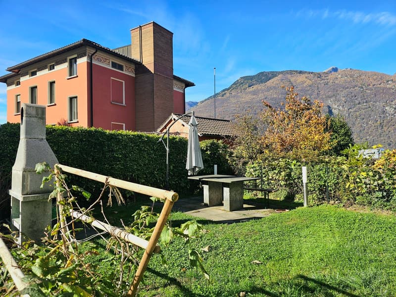a table with a closed umbrella, stone fireplace, stone bench, grass, mountain range in the background