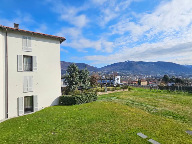 Building with white walls, red roof, multiple windows, surrounded by green lawn, mountains in the background