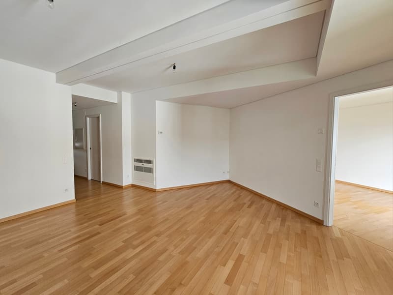 Large, empty room, wooden floor, white walls, two doorways, power outlets, AC unit in one corner