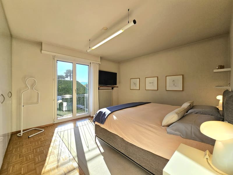 Large bed with pillows, sliding glass door leading to balcony, television, radiator, picture frames, wardrobe, side table with lamp, hanging ceiling light