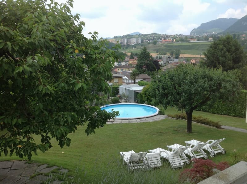 Garden with a round swimming pool, lounge chairs, trees, grass, and scenic view of the town and mountains