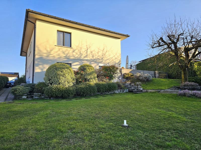 2-story house, light yellow exterior, white windows, shrubs and trees, concrete pathway, concrete driveway, car parked, garden, clear blue sky