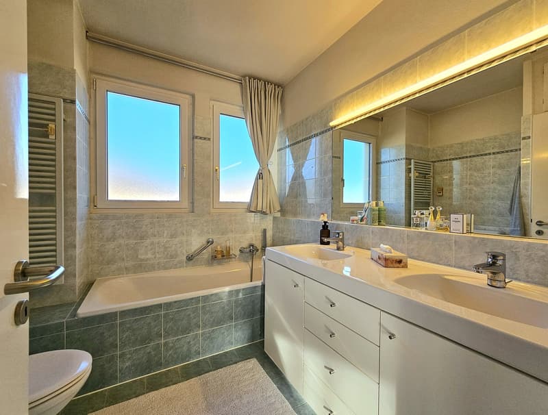 large bathtub, twin sinks, spacious mirror, tiled floor, large windows, towel hanging on the wall