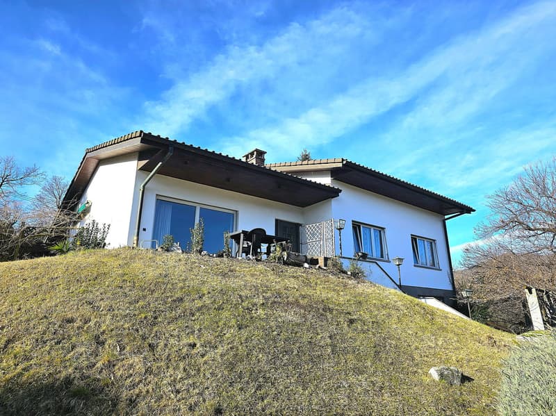 white villa on a raised ground floor, brown roof, glass doors and windows, a table on the porch, trees in the distance