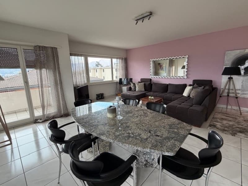 Modern furnished room with a dining table, chairs, a couch, a lamp, a mirror, and a rug. The room has a view of the outside.