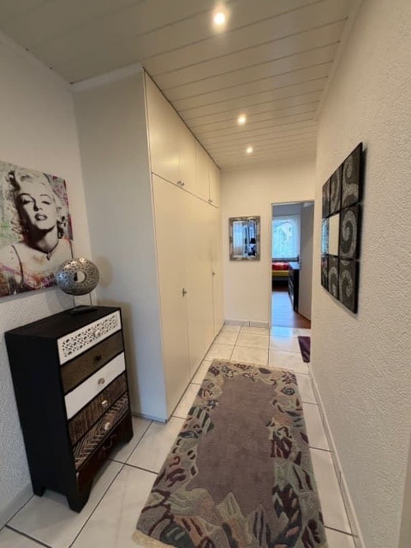 A hallway with white tiles, a rug, a dresser, and a cabinet. There are paintings on the wall and a mirror.